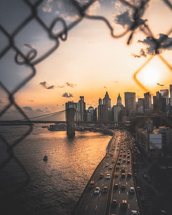 Captivating view of New York City skyline with Brooklyn Bridge during sunset, framed artistically by chain-link fence. HD phone wallpaper