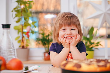 A happy young girl smiling indoors surrounded by plants and a pastry, with natural light streaming in. HD wallpaper