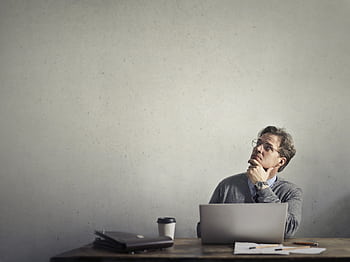 Thoughtful businessman looking up while working at his desk with a laptop and coffee. HD wallpaper