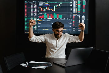 Businessman celebrates stock market success with hands raised in excitement at a trading desk. HD wallpaper