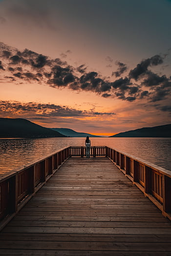 A tranquil sunset view from a wooden pier overlooking a calm lake and distant mountains. HD phone wallpaper