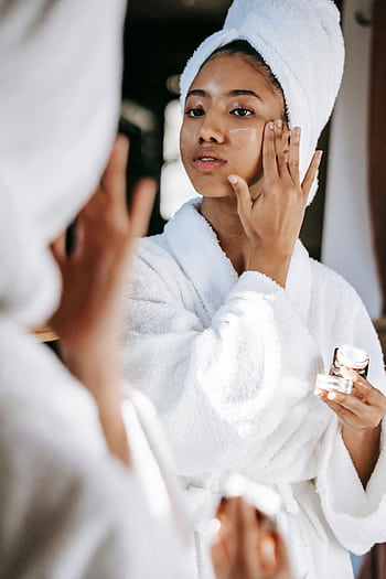 A young woman in a bathrobe applies skincare cream in a bathroom mirror, promoting wellness and beauty. HD phone wallpaper