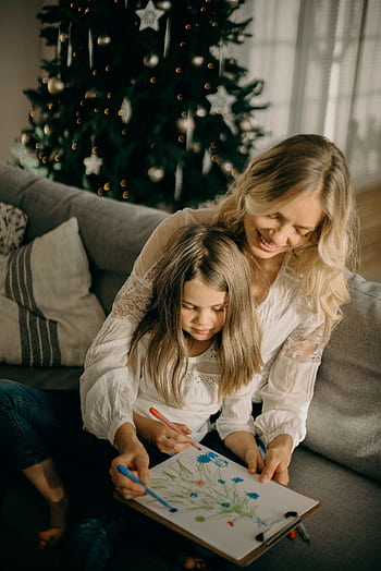A mother and daughter enjoy drawing together by a Christmas tree, capturing a cozy holiday moment indoors. HD phone wallpaper