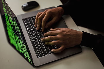 Close-up of hands typing code on a laptop with green text on screen, suggesting hacking or programming. HD wallpaper