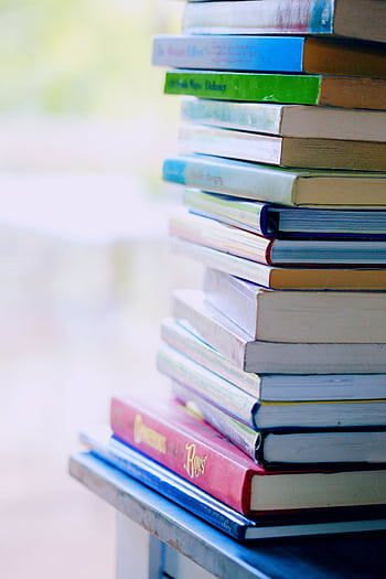 A tall stack of colorful hardcover books on a table indoors, perfect for library-themed concepts. HD phone wallpaper