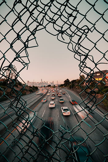 Traffic flows on a highway framed by a broken fence with San Francisco cityscape at sunset. HD phone wallpaper