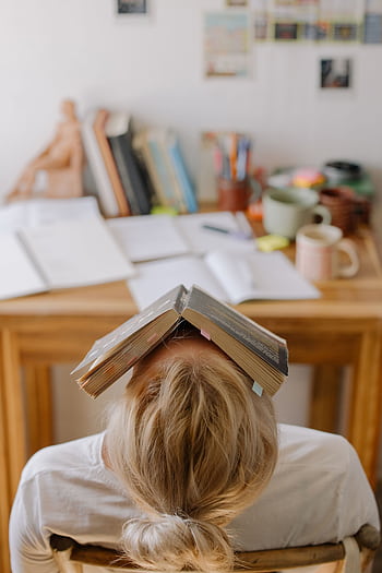 Student feeling stress and exhaustion while studying at a cluttered desk with an open book on their head. HD phone wallpaper