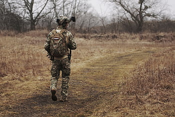 A soldier in camouflage walking along a grassy path through a forested area. HD wallpaper