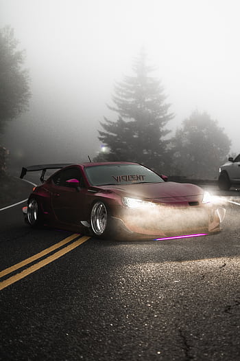 Red sports car with headlights on, driving through a foggy mountain road with pine trees. HD phone wallpaper