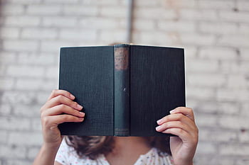 A woman holds an open book, reading in front of a brick wall. HD wallpaper