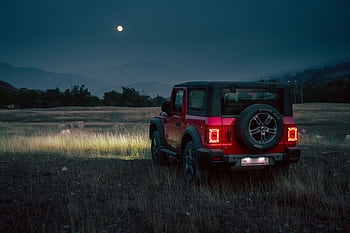 A red Mahindra Thar SUV sits in a grassy field under the moonlight in rural India. HD wallpaper