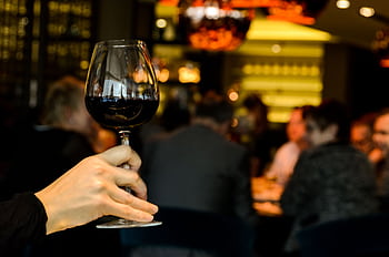 A hand holding a glass of red wine with a blurred group dining in a stylish restaurant. HD wallpaper