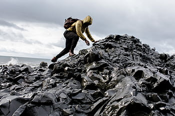 Person in yellow jacket climbs black rocky terrain by the ocean under cloudy skies. HD wallpaper