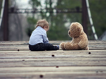 Adorable toddler sitting with a teddy bear on a wooden bridge, enjoying a peaceful moment outdoors. HD wallpaper