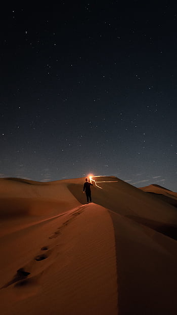 A lone figure walks across desert dunes beneath a starry night sky, the light guiding their way. Perfect for nature and adventure themes. HD phone wallpaper