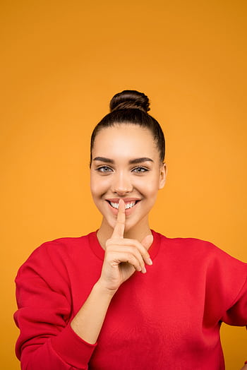 Young woman smiling with finger on lips against a vivid yellow background. HD phone wallpaper