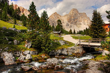 A beautiful mountain landscape with a flowing stream, lush greenery, and a bridge under a cloudy sky. HD wallpaper
