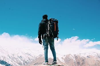 A lone hiker with a backpack surveys snow-capped mountains under a clear blue sky. HD wallpaper