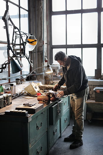 Male artisan focusing on metalwork in a rustic industrial workshop. Indoor scene with tools. HD phone wallpaper