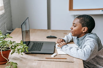 A young boy focused on online learning at home, using a laptop and notebook, illustrating modern education. HD wallpaper