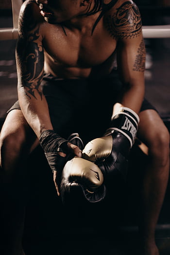 A muscular boxer with tattoos sitting shirtless in a gym holding boxing gloves. HD phone wallpaper