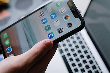 Person holding a smart in front of a laptop showcasing various app icons, highlighting digital connectivity. HD wallpaper