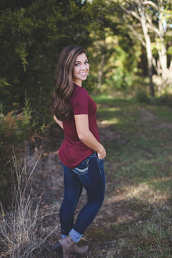 A young woman with long hair smiles outdoors in a forest setting, wearing casual attire. HD phone wallpaper
