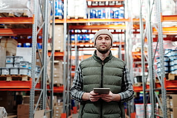 A warehouse worker wearing a beanie uses a tablet to manage inventory in a storeroom with shelves. HD wallpaper