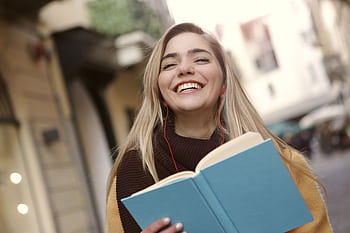 Smiling woman enjoying a book with earbuds in an urban outdoor setting. HD wallpaper