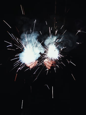 Close-up of hands holding bright sparklers emitting smoke and sparks against a dark background. HD phone wallpaper