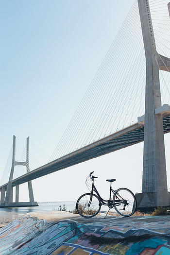 A bicycle with vibrant graffiti beneath a striking suspension bridge under a clear blue sky. HD phone wallpaper