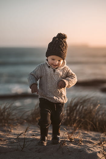Adorable toddler in warm knitwear exploring a sandy beach with a joyful smile at sunset. HD phone wallpaper