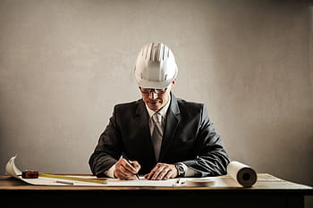 Engineer in a suit drafting architectural plans at a desk indoors. HD wallpaper