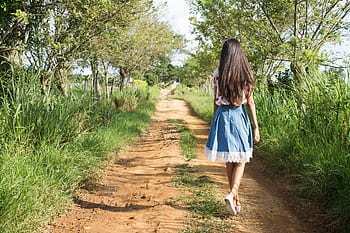 A woman in a blue skirt walks down a rural dirt path surrounded by lush greenery. HD wallpaper