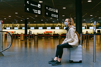 A woman wearing a face mask sits on luggage in an airport terminal amid the pandemic. HD wallpaper
