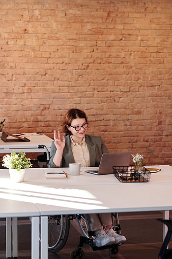 A cheerful businesswoman in a wheelchair working remotely from a modern office setting. HD phone wallpaper