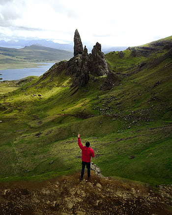 A lone hiker in a red jacket stands before the iconic Old Man of Storr on the Isle of Skye, Scotland. HD phone wallpaper