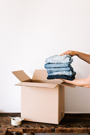 Person packing folded jeans into a cardboard box, preparing for a move or organization task. HD phone wallpaper