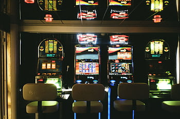 Slot machines in a dimly lit casino with reflective ceiling and empty chairs. HD wallpaper