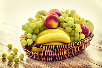 A colorful wicker basket filled with fresh grapes, bananas, and apples on a wooden table. HD wallpaper