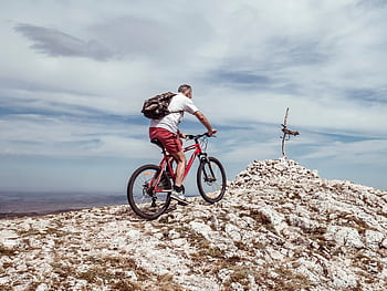 An elderly man biking uphill on a rocky mountain trail under a cloudy blue sky. HD wallpaper
