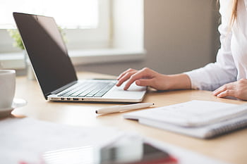 A businesswoman typing on a laptop at a bright indoor workspace with notebooks and a pen. HD wallpaper