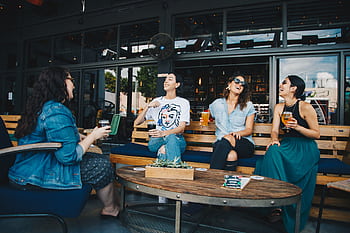 Four friends laughing and enjoying drinks together at an outdoor cafe setting, reflecting happiness and camaraderie. HD wallpaper