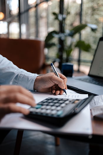 A person calculating finances with a calculator and pen on a desk indoors. HD phone wallpaper