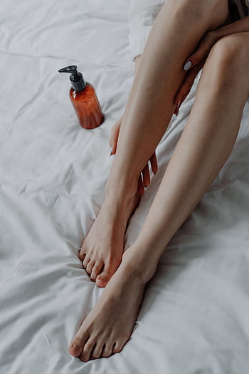 A young woman applies cream on her legs, sitting on a white bed at home. HD phone wallpaper