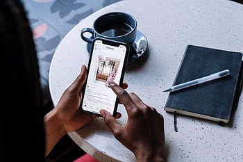 Hands holding smart at a cafe table with coffee and a notebook, focusing on social media browsing. HD wallpaper