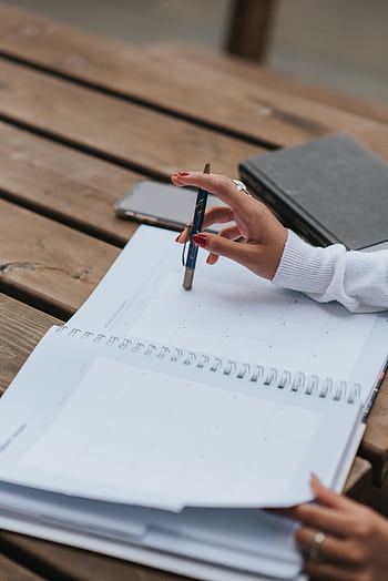 A woman's hand writes in a spiral notebook on a wooden table outdoors. HD phone wallpaper