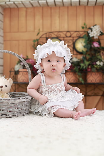 Cute baby in a lace dress and hat sitting indoors with a teddy bear basket, floral background. HD phone wallpaper