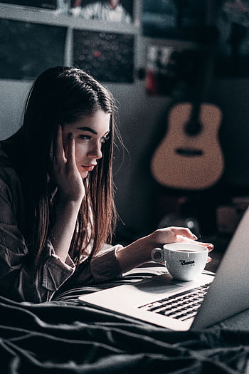 Woman lying on bed, focused on laptop with a coffee cup in hand, embracing remote work lifestyle. HD phone wallpaper