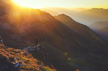 Lone hiker standing on a rocky mountain range at sunrise, basking in golden sunlight. HD wallpaper
